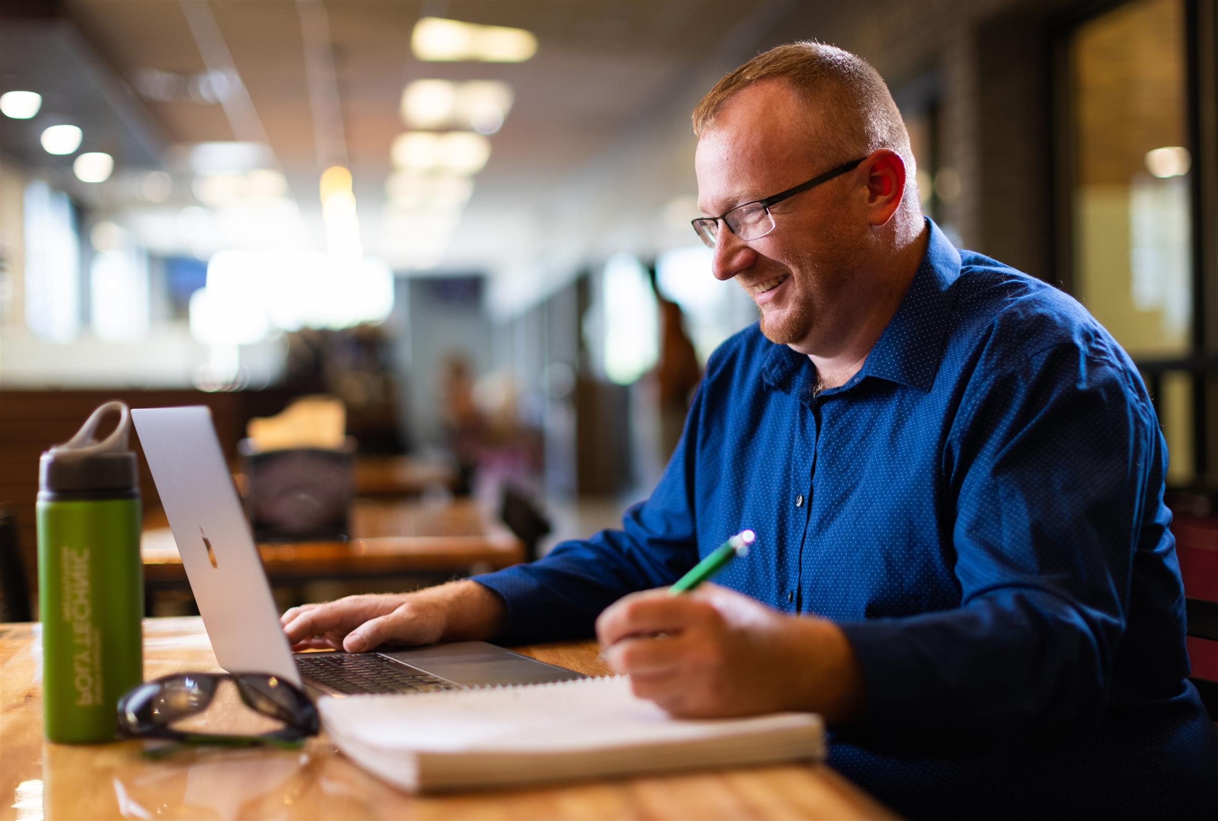 一个穿着蓝色衬衫，戴着眼镜的男人坐在咖啡店的桌子旁做作业. 桌子上有一台笔记本电脑，笔记本，太阳镜和一个水瓶. 